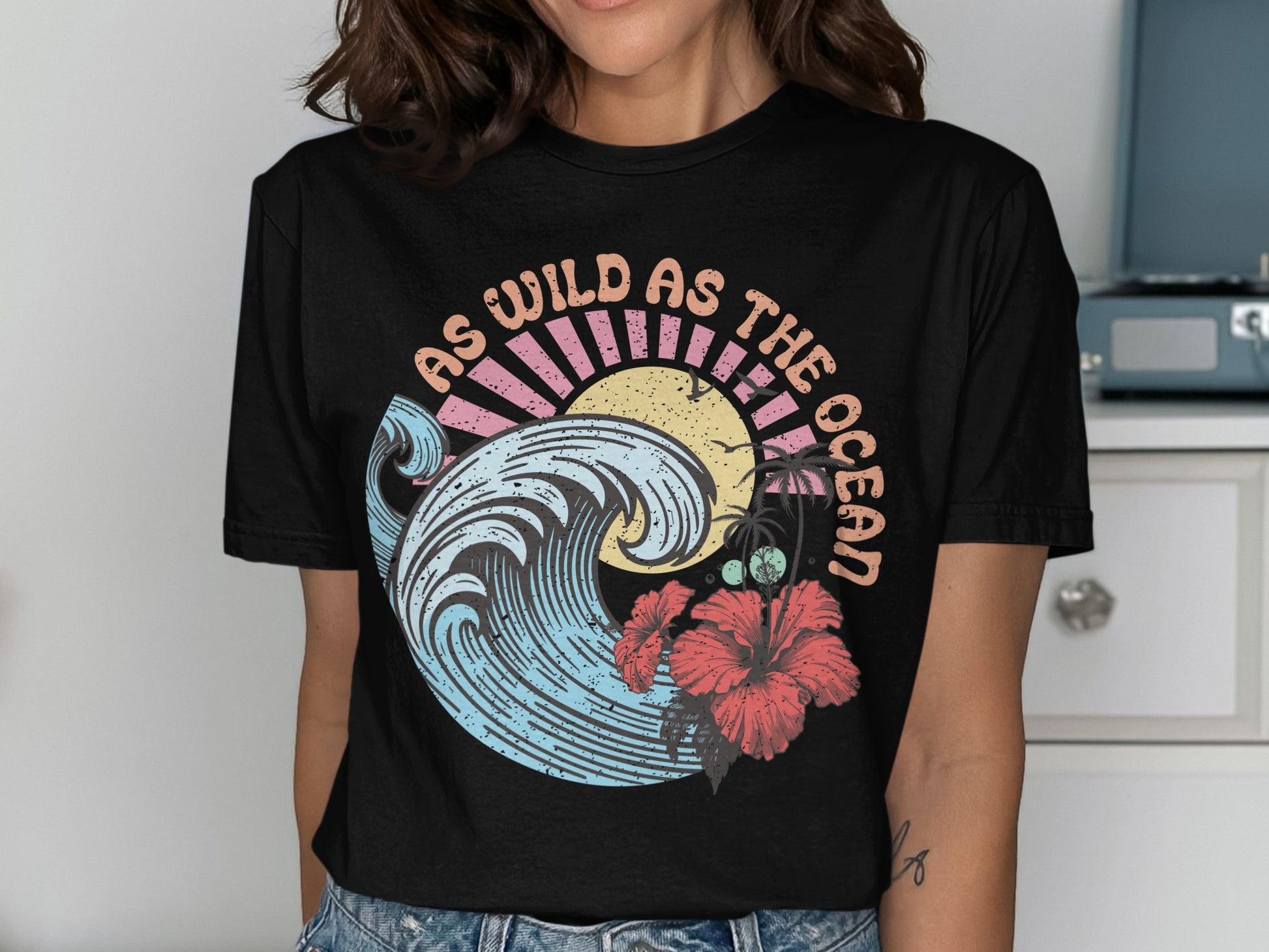 Black t-shirt with a graphic featuring a large wave, hibiscus flower, sun, and text "AS WILD AS THE OCEAN".