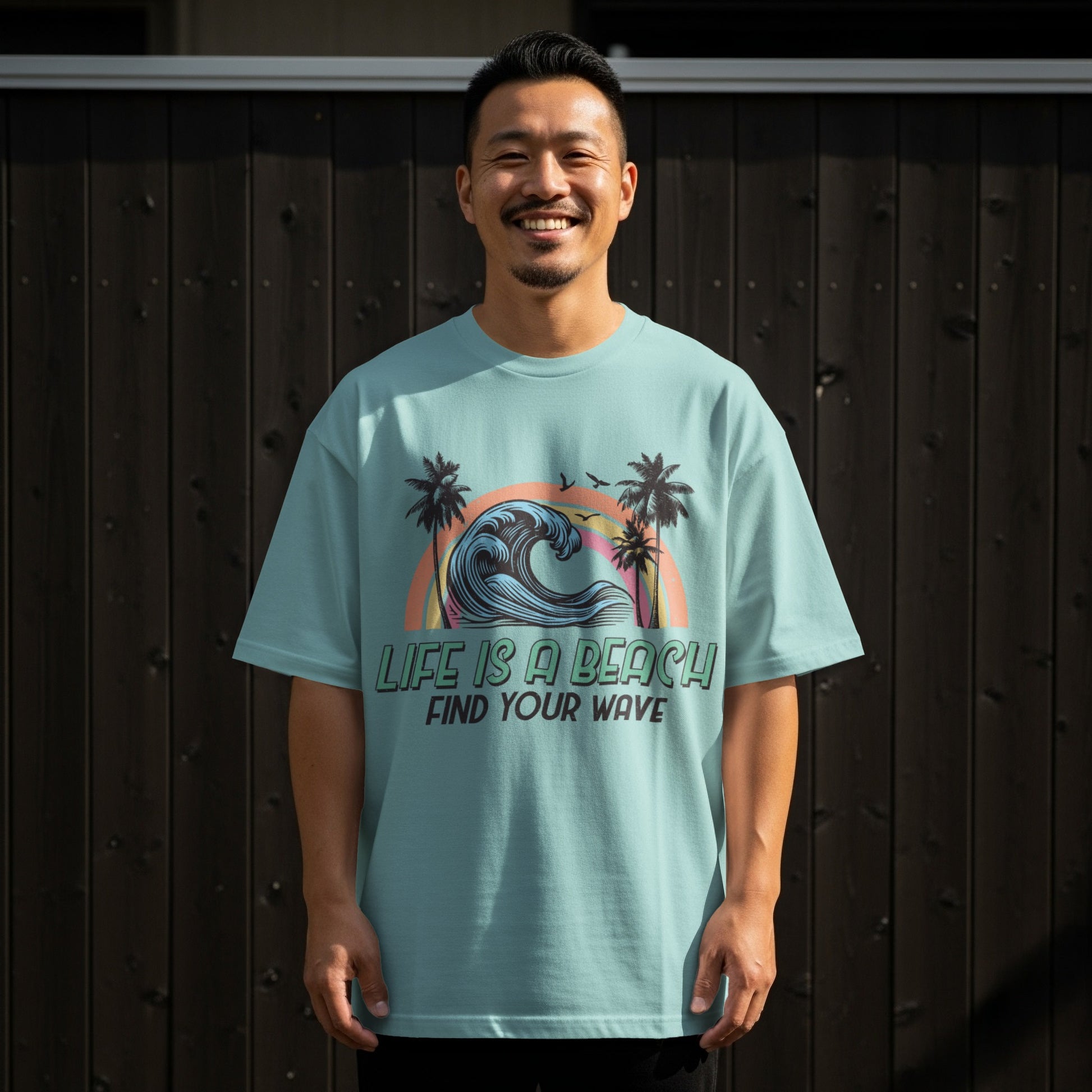Man wearing a teal t-shirt with a graphic of a rainbow wave and palm trees.
